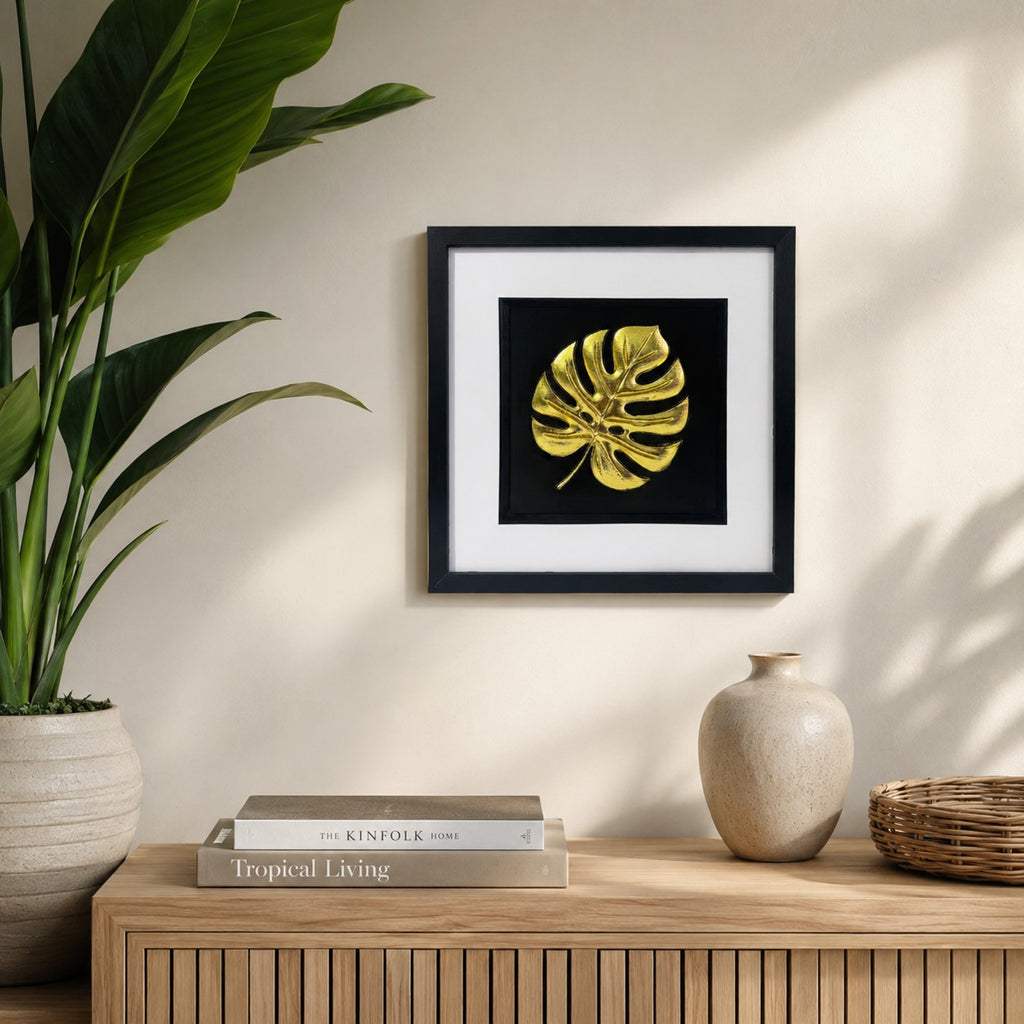 Gold monstera leaf wall art displayed in a tropical inspired interior, featuring a 33 cm square framed hand gilded gold leaf 3D relief botanical leaf design on a black background.