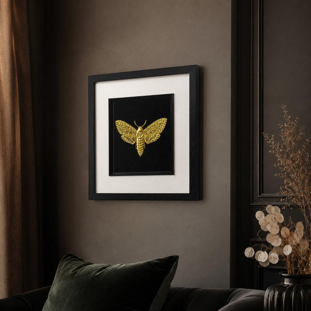 55 degree angled view of framed gold moth wall art in a black square frame with white mount, hung on a moody wall in a stylish dark interior.