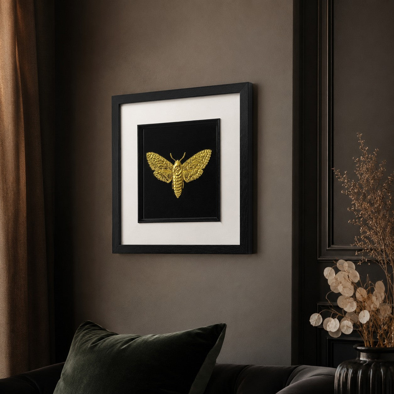 55 degree angled view of framed gold moth wall art in a black square frame with white mount, hung on a moody wall in a stylish dark interior.