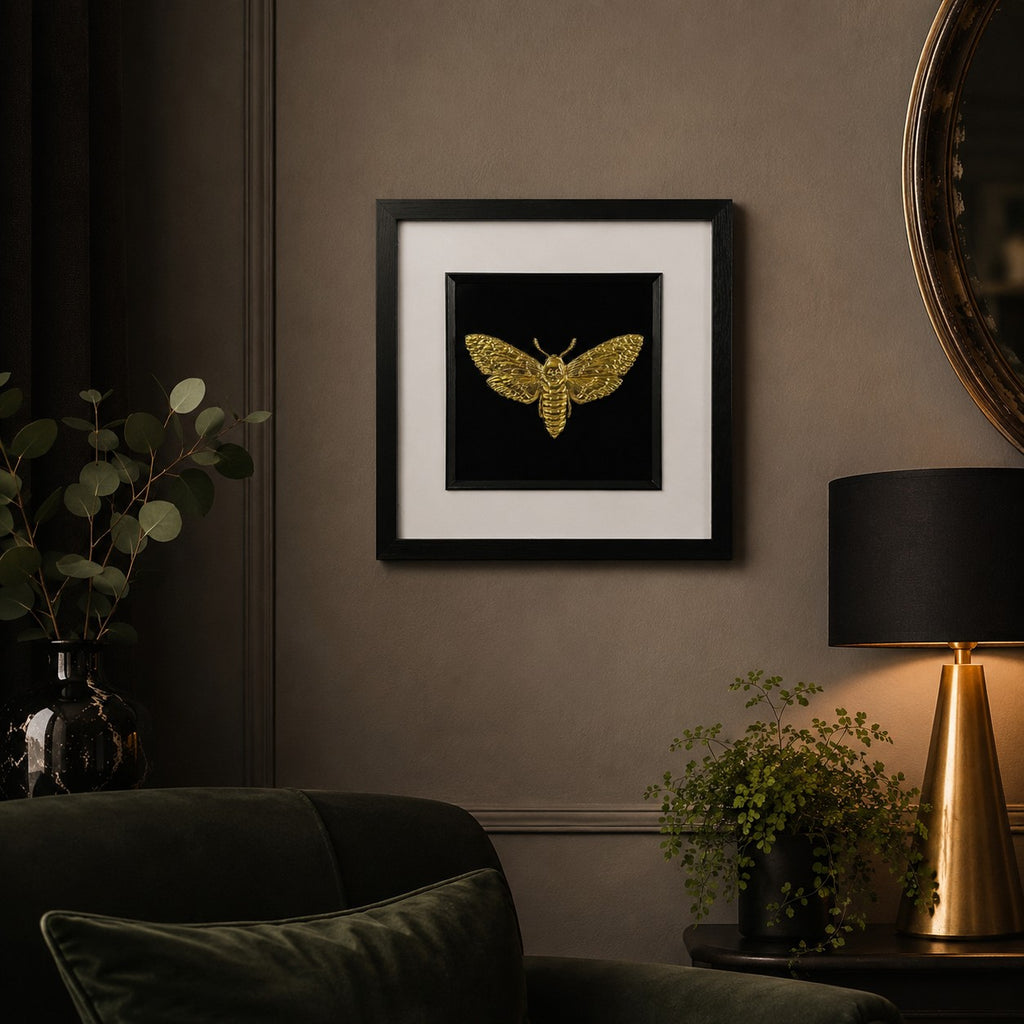 Framed gold moth wall art in a black square frame with white mount, displayed on a moody taupe wall in an elegant living room with dark furniture and warm lighting.
