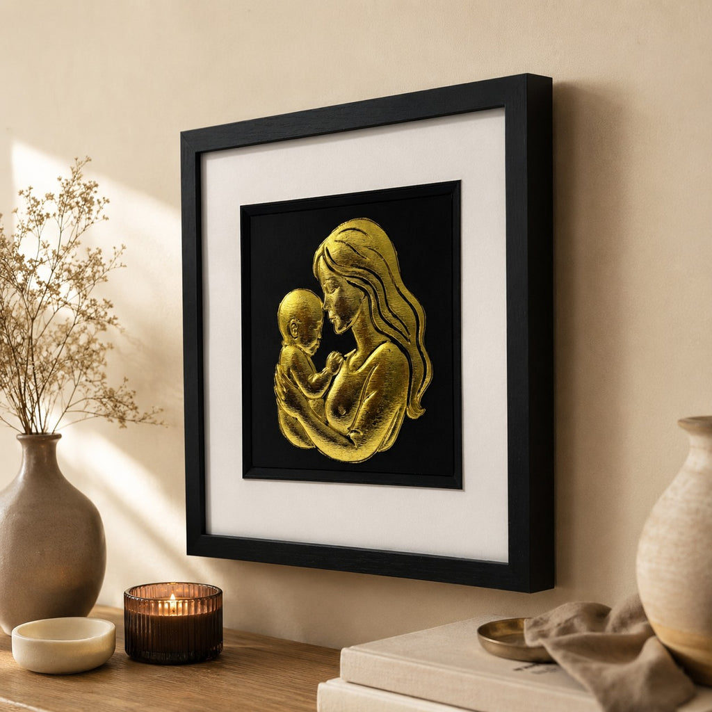 Angled lifestyle view of gold mother and baby wall art in a black square frame with white mount, displayed with warm neutral decor, ceramic vases, books and a lit candle.