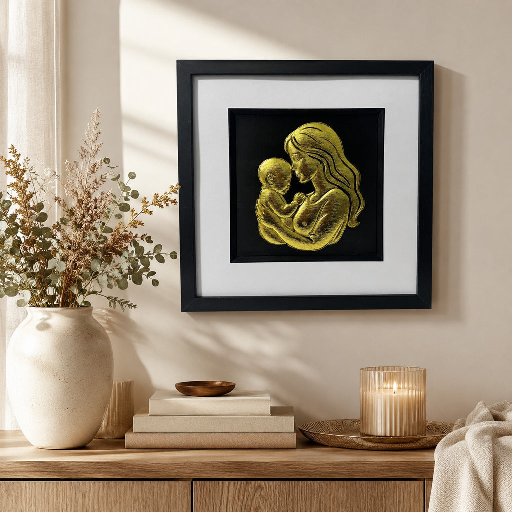 Gold mother and baby wall art in a black frame with white mount, styled on a neutral wall above a light wood console with soft dried florals, books and candlelight.