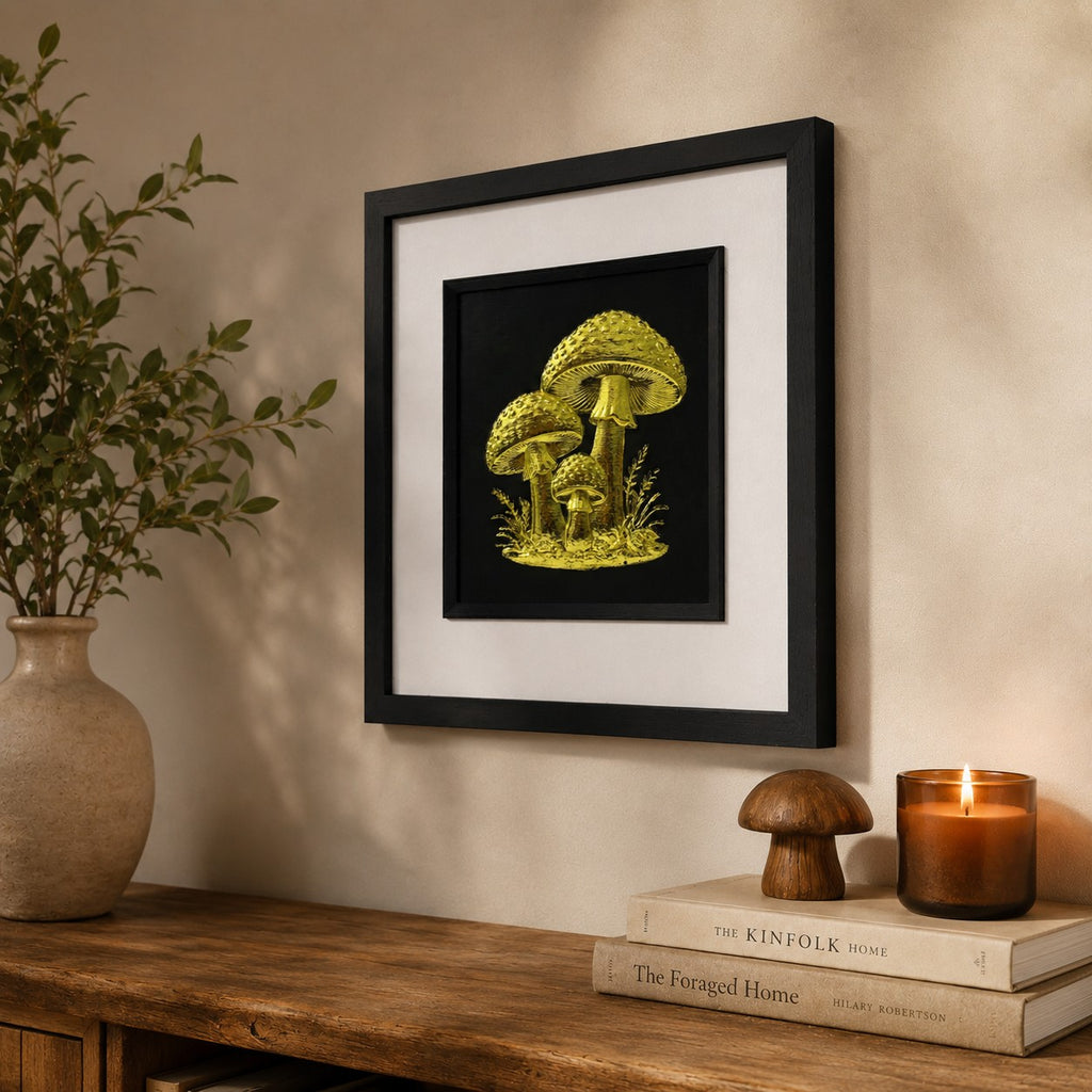 Angled lifestyle view of gold mushroom wall art in a black square frame with white mount, displayed in a warm rustic interior with botanical decor, books and a lit candle.