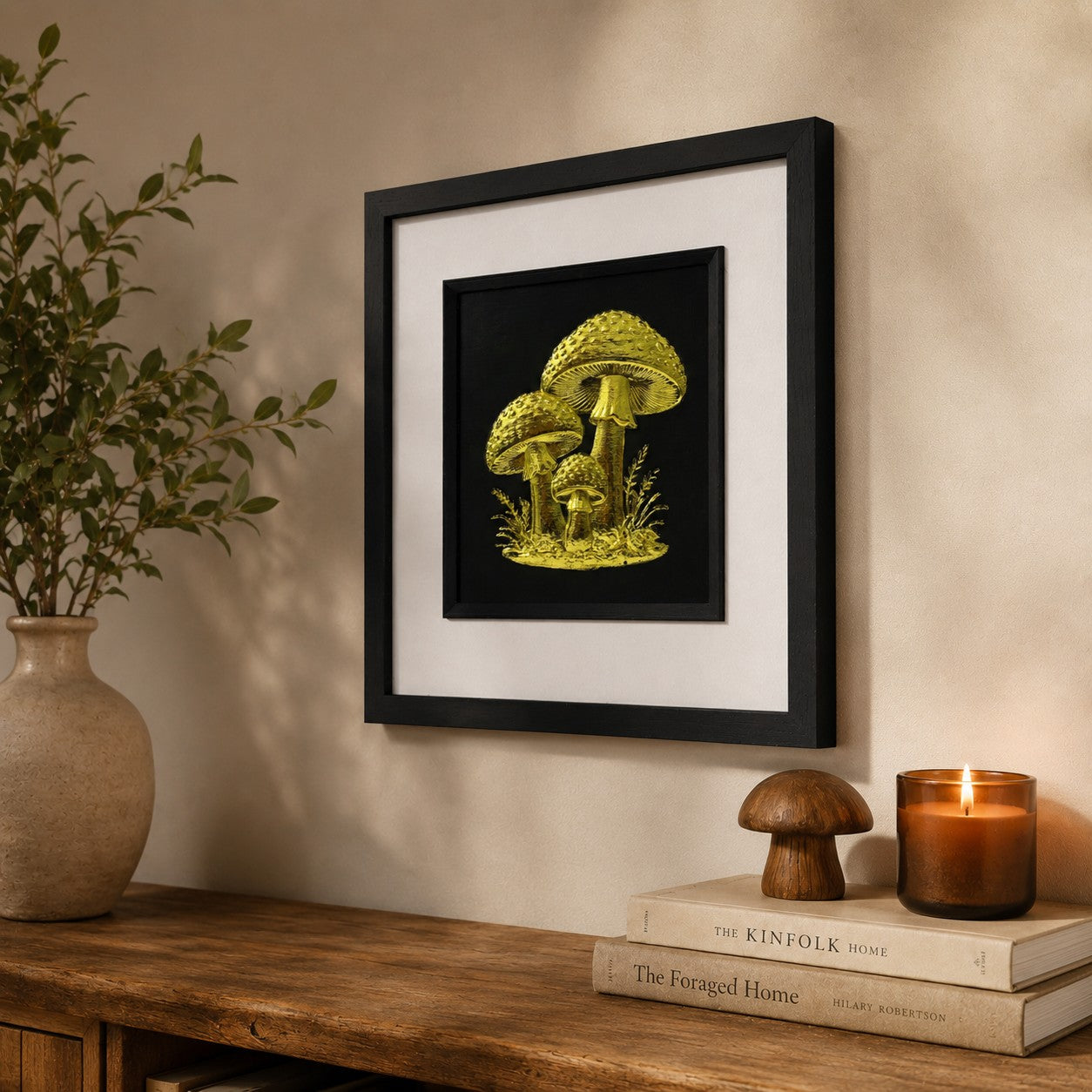 Angled lifestyle view of gold mushroom wall art in a black square frame with white mount, displayed in a warm rustic interior with botanical decor, books and a lit candle.