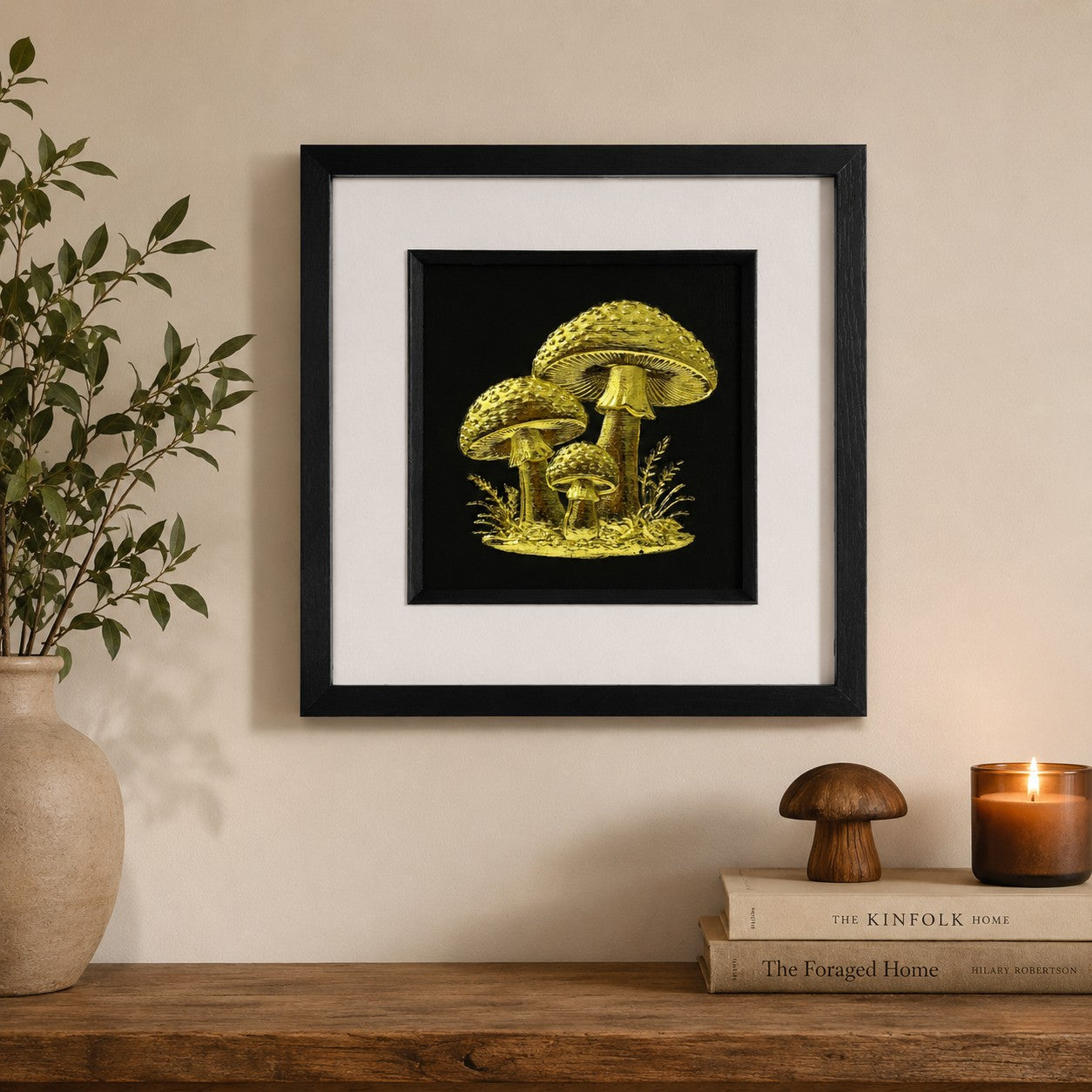 Gold mushroom wall art in a black frame with white mount, styled on a neutral wall above a rustic wooden console with leafy stems, books, candlelight and natural decor.