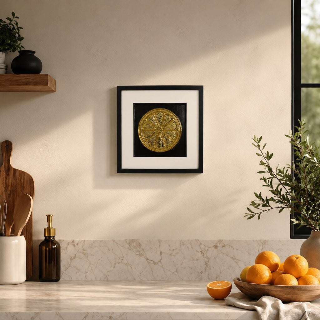 Gold 3D orange slice artwork in black frame styled on a kitchen wall with oranges, greenery and marble-style worktop.