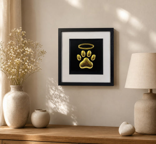 Gold paw print memorial wall art with halo displayed in a 33cm square black frame on a warm neutral wall above a wooden console with soft remembrance decor.