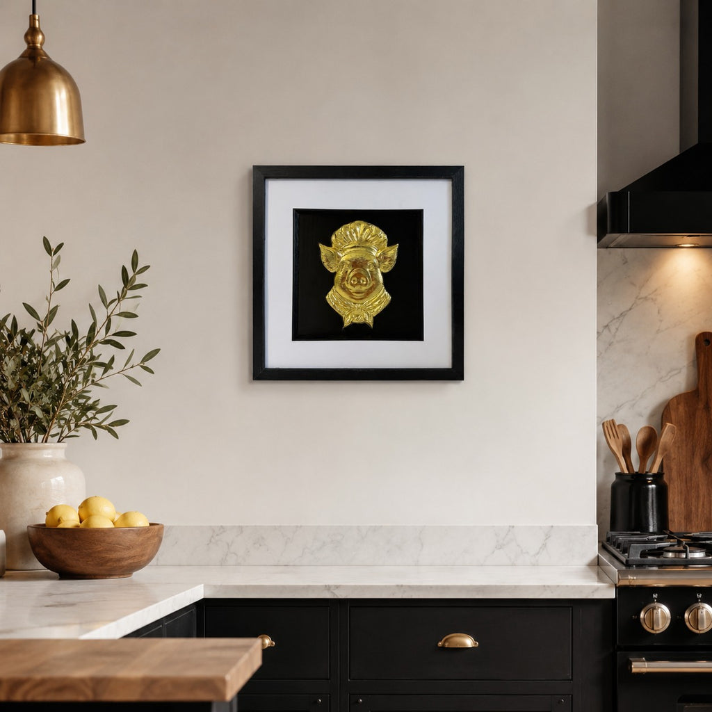 Gold pig chef framed artwork hanging in a modern kitchen with brass pendant light, marble worktop and black cabinetry