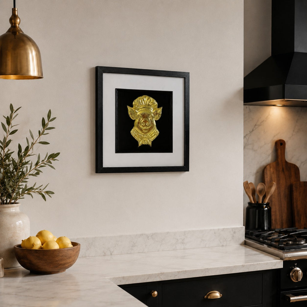 Angled view of gold pig chef framed kitchen artwork on a neutral wall beside black stove hood and wooden utensils