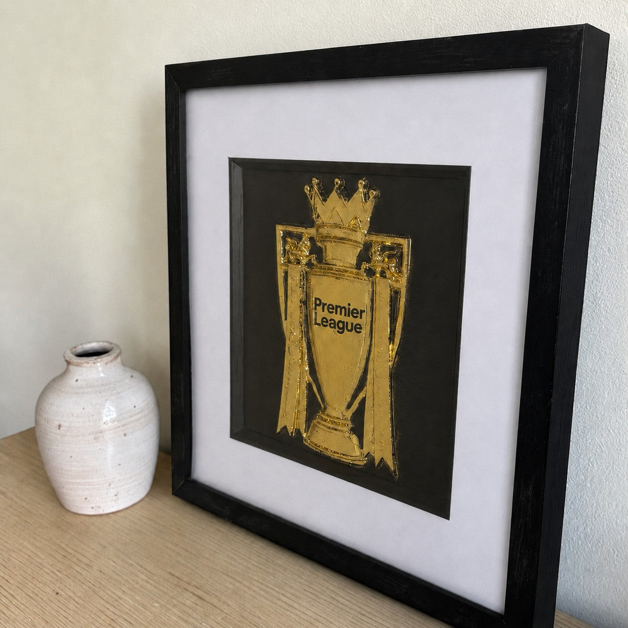 Gold Premier League trophy framed artwork styled on a wooden shelf beside a small ceramic vase