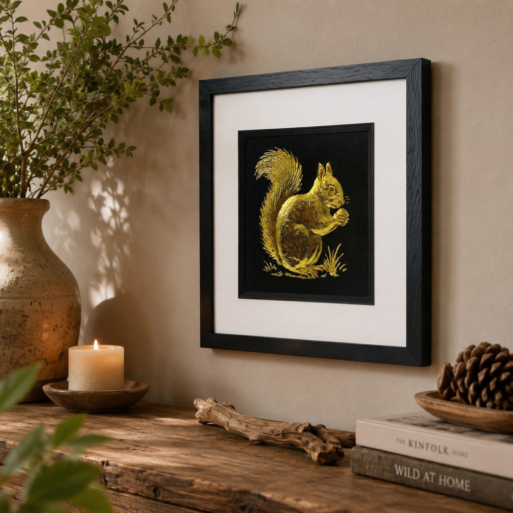 Angled lifestyle view of gold squirrel wall art in a black square frame with white mount, displayed with rustic natural decor, a ceramic vase, candle, books and pinecone accents.