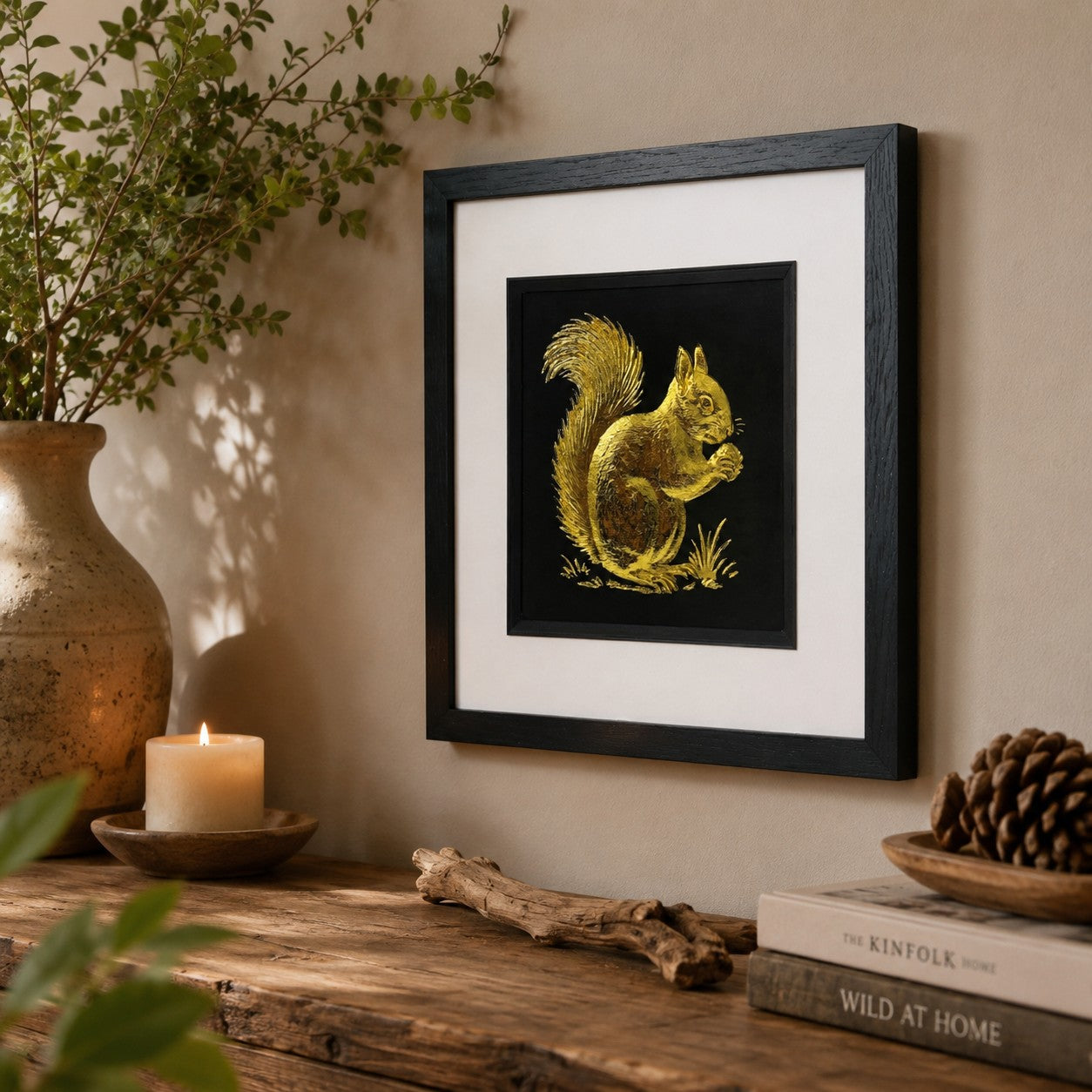 Angled lifestyle view of gold squirrel wall art in a black square frame with white mount, displayed with rustic natural decor, a ceramic vase, candle, books and pinecone accents.