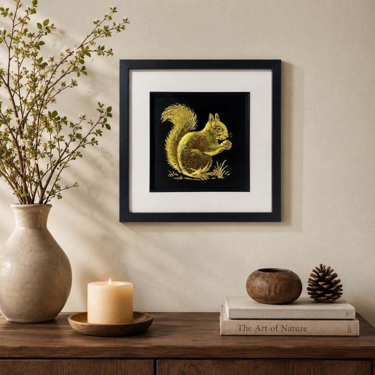 Gold squirrel wall art in a black frame with white mount, styled on a neutral wall above a dark wood console with a ceramic vase, candle, books and natural decor accents.