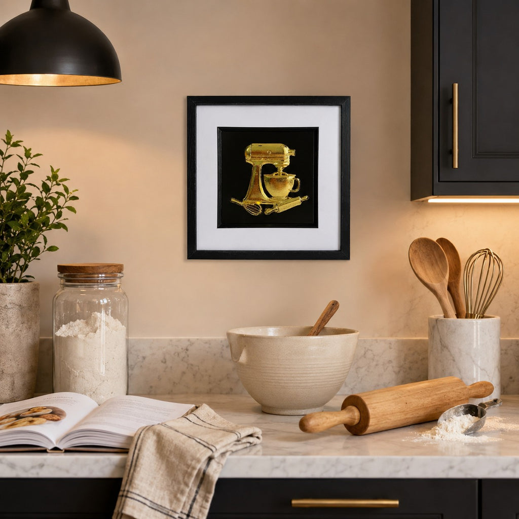 Gold stand mixer relief artwork in a black square frame styled on a kitchen wall above a baking worktop.