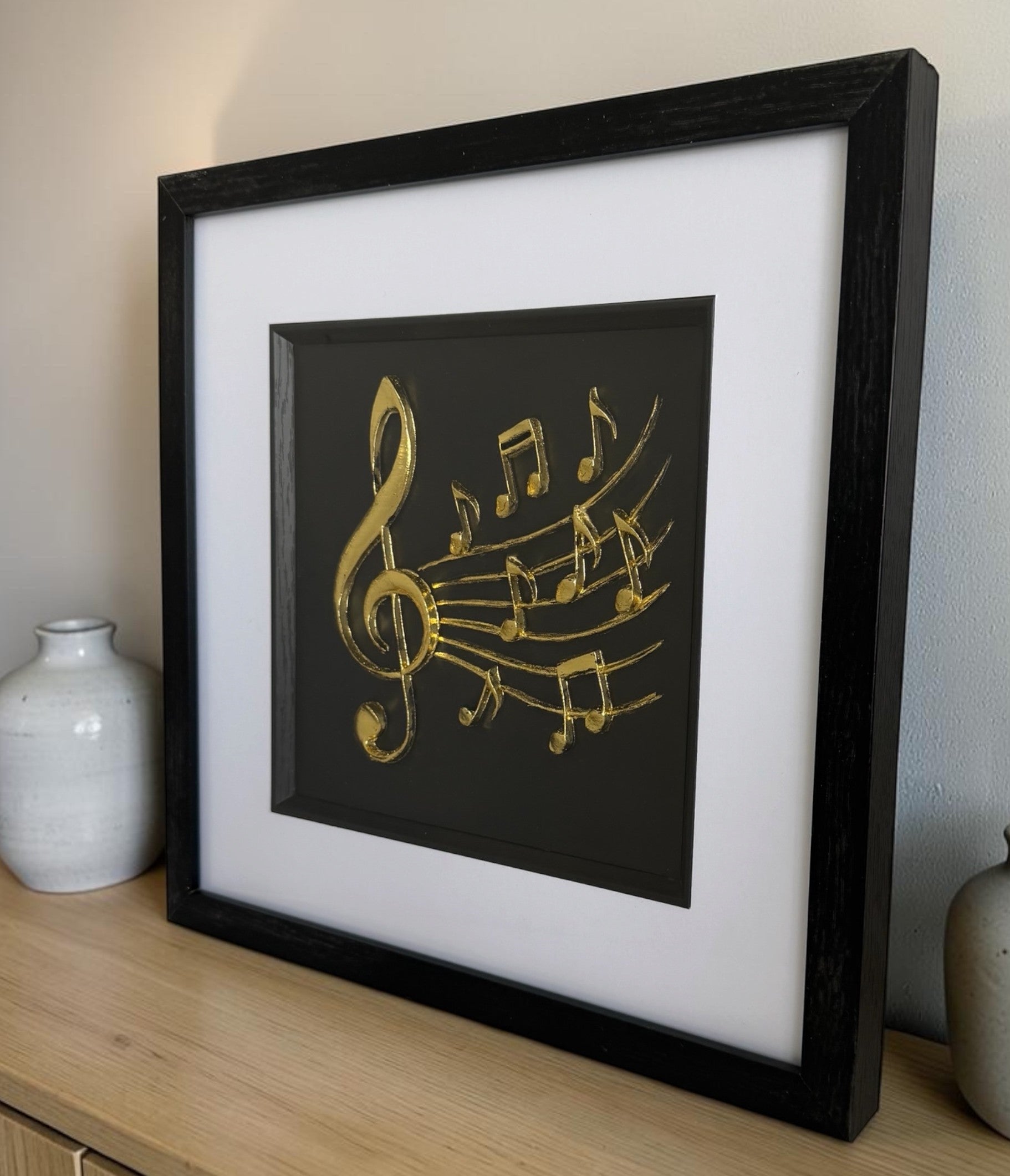 Gold treble clef and music notes relief in a black frame with glass front, styled on a pale wood sideboard