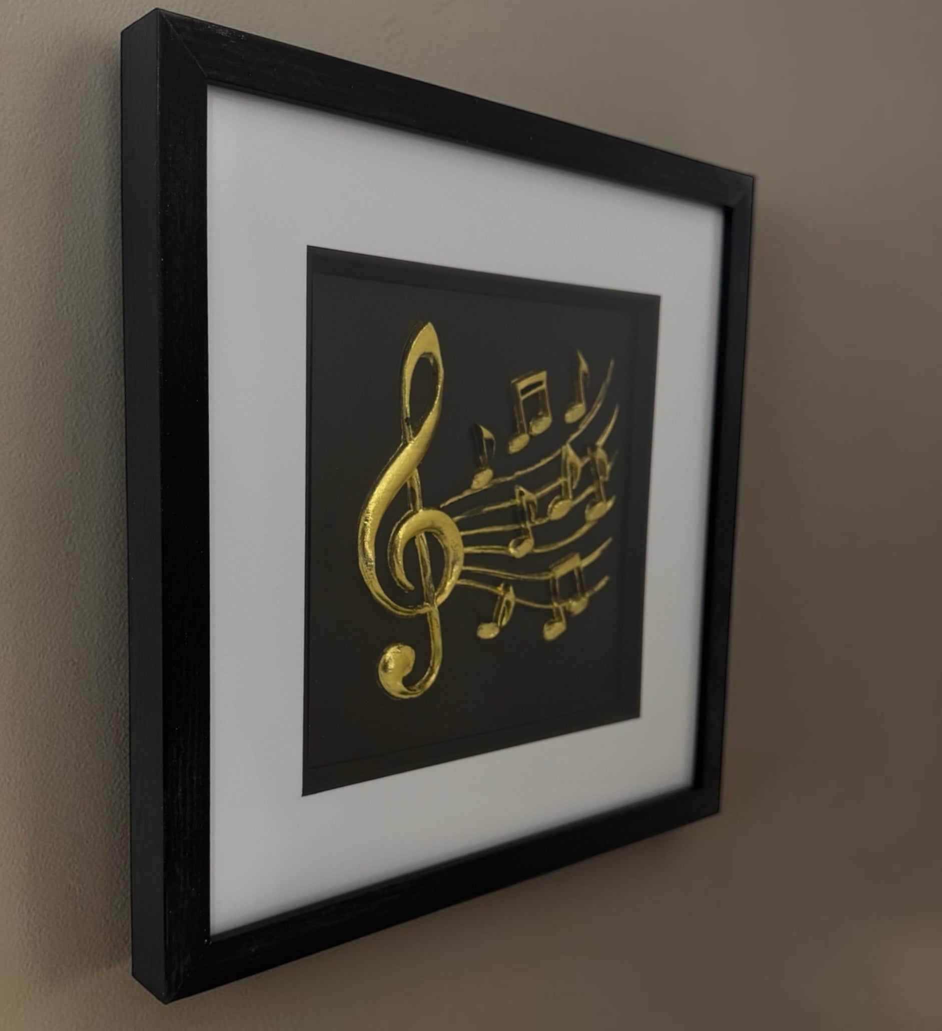 Gold treble clef and music notes framed artwork hanging on a neutral wall, showing the black box frame depth