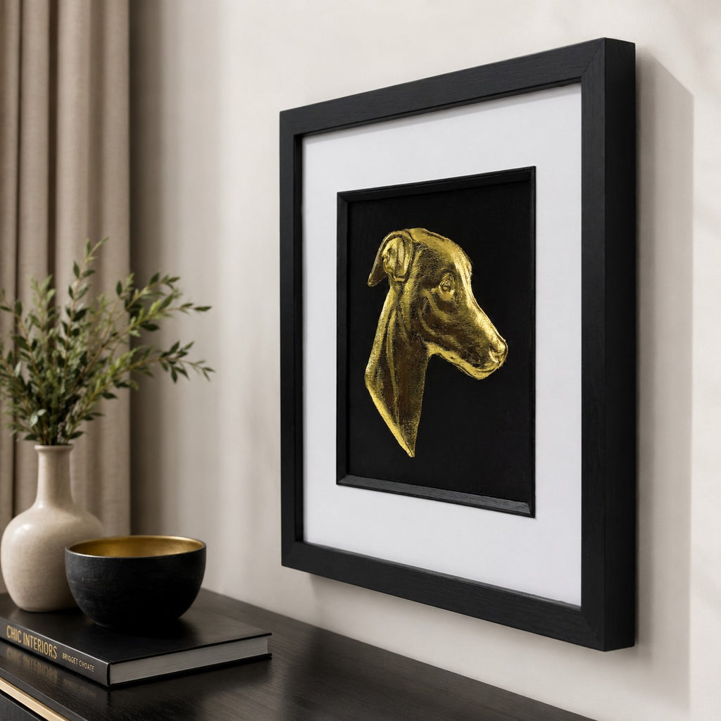 Angled lifestyle view of gold whippet wall art in a black square frame with white mount, displayed on a neutral wall with elegant black, gold and natural decor accents.