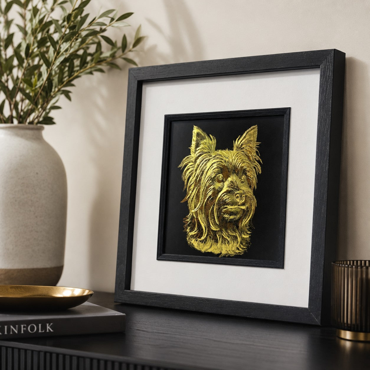 Angled lifestyle view of gold Yorkshire Terrier wall art in a black square frame with white mount, displayed with neutral decor, a ceramic vase, gold tray and dark console table.