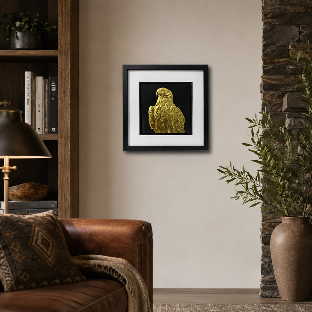 Framed golden eagle 3D wall art with gold relief detail, displayed on a neutral wall in a warm rustic living room with leather, wood and natural decor.
