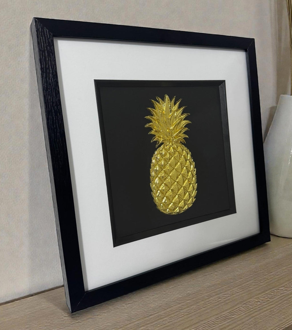Gold leaf pineapple raised relief artwork in black frame with white mount, shown on a desk against a neutral wall in natural light