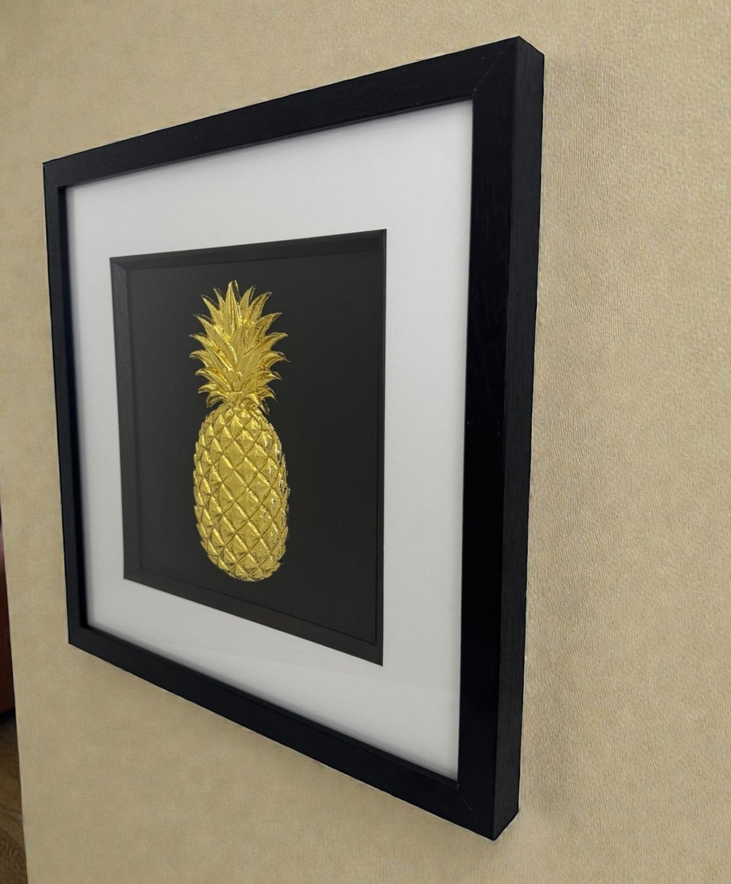 Gold leaf pineapple raised relief artwork in black frame with white mount, shown on a desk against a neutral wall in natural light