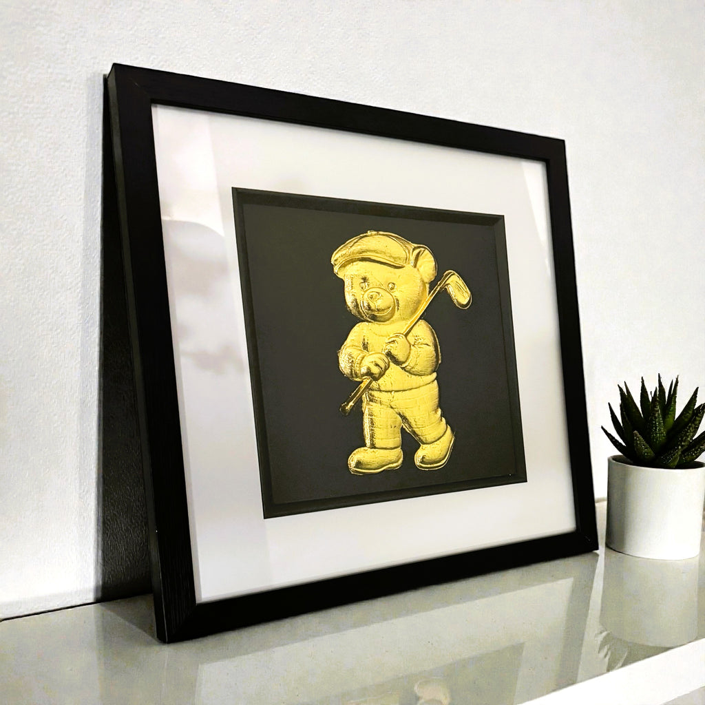 Framed gold teddy bear golfer art displayed on white nursery shelf.