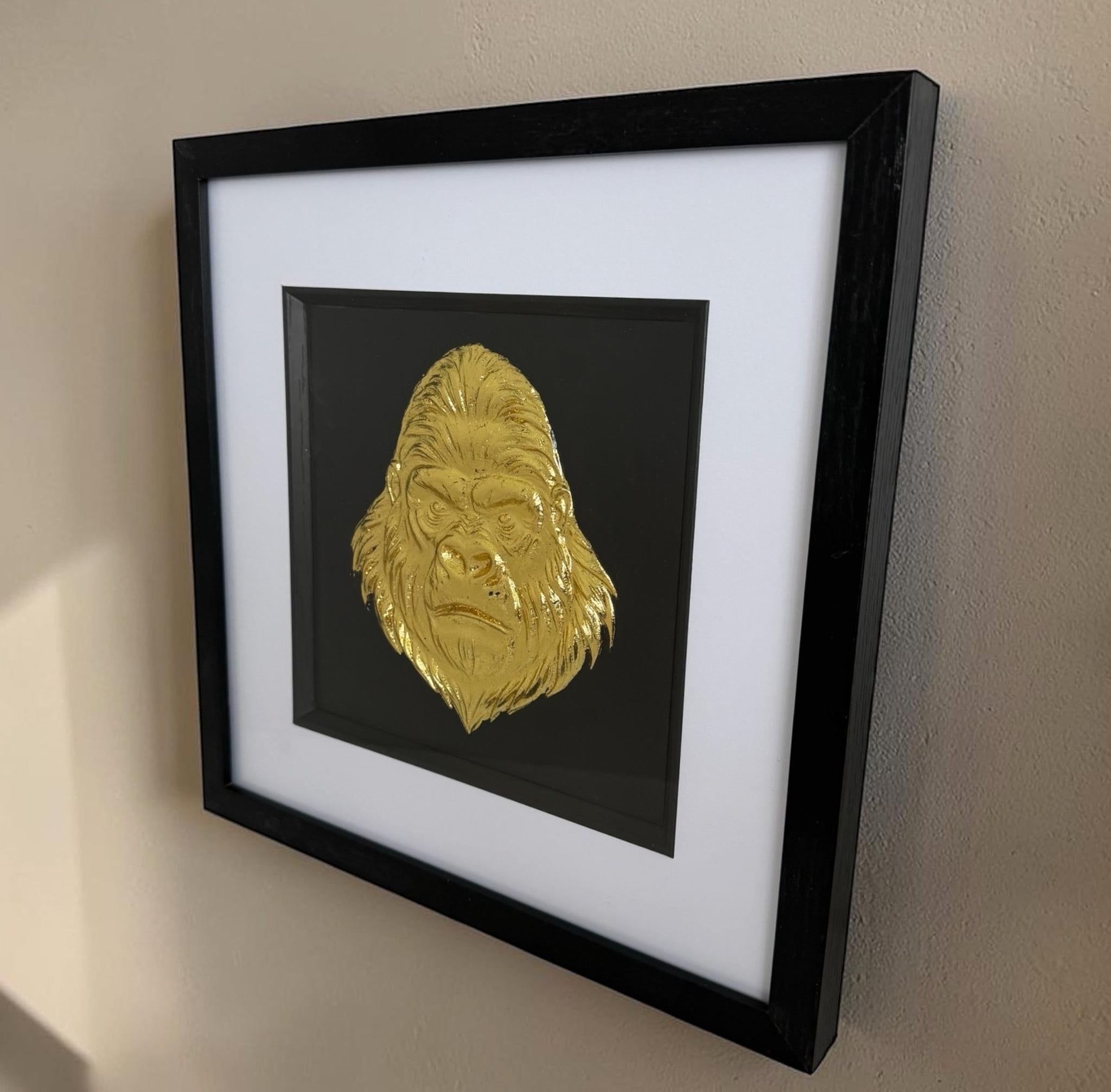 Angled gold gorilla head relief artwork with black centre, white mount and black square frame.