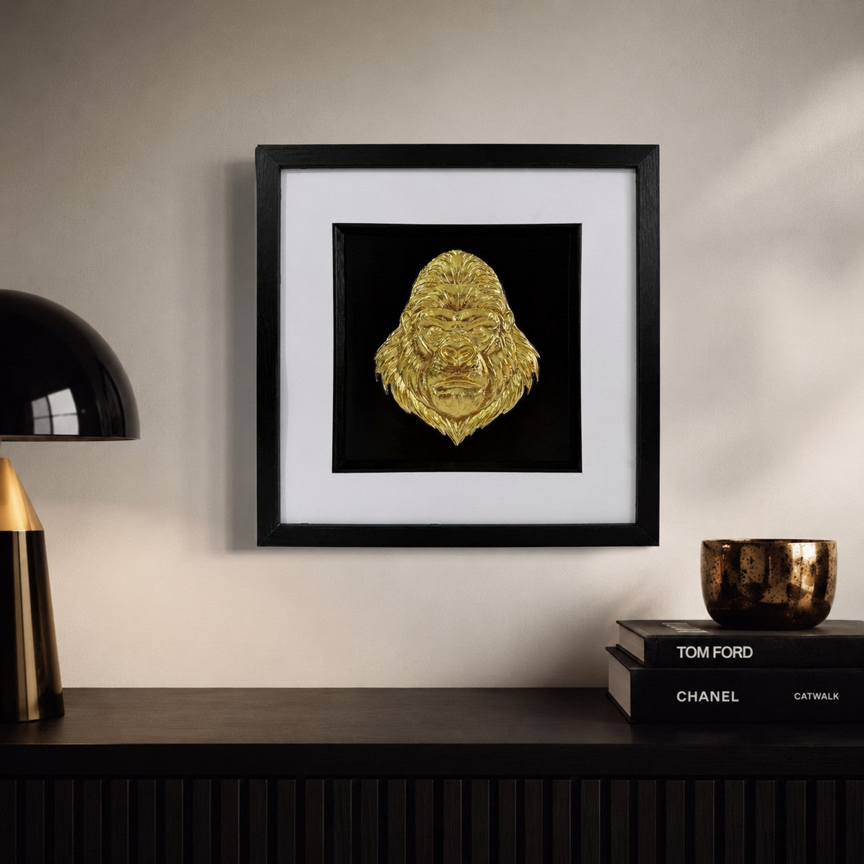 Gold gorilla head artwork styled above a dark sideboard with books, lamp and warm neutral decor.