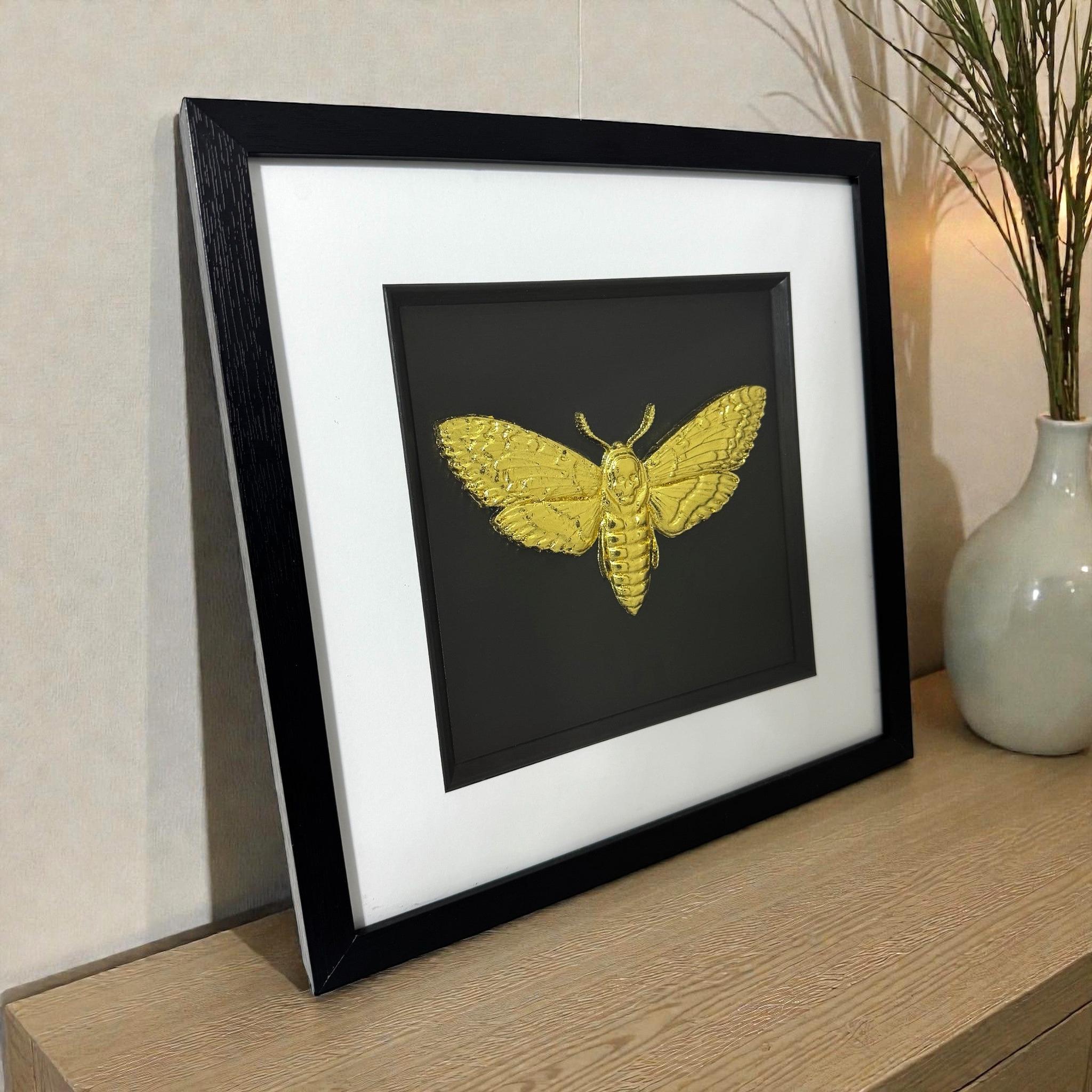 Death’s head hawkmoth 3D wall art in gold leaf, framed in a black frame with white mount, styled on a wooden surface with a vase.