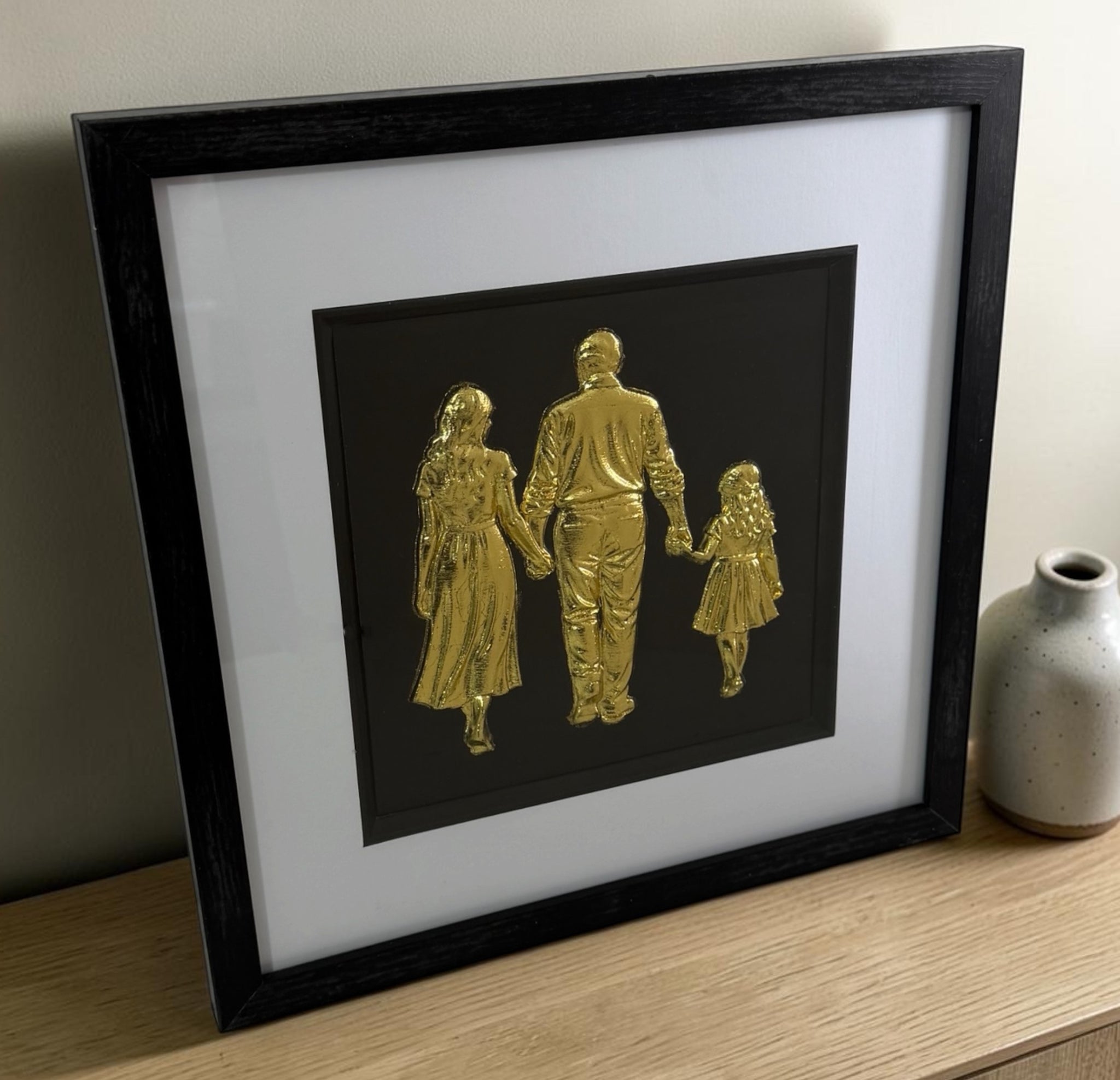 Angled tabletop view of Gold family relief artwork with glass front, black frame and white mount.