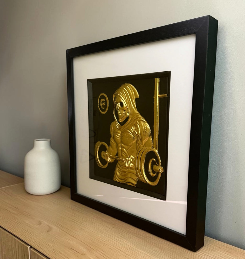 Gold grim reaper lifting weights framed wall art leaning on an oak sideboard beside a white vase, angled to show the raised relief through the glass