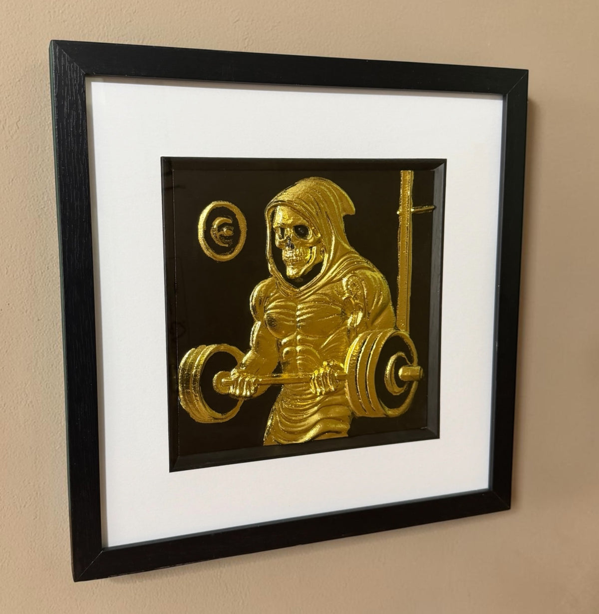 Gold grim reaper gym 3D wall art hanging on a beige textured wall, viewed from the side to show the depth of the black box frame and the raised barbell
