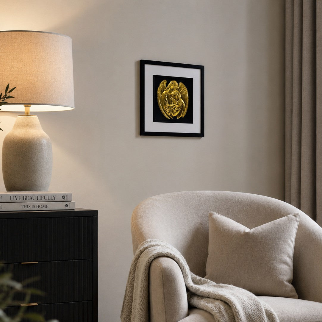 Angled lifestyle view of framed gold leaf guardian angel wall art displayed on a living room wall beside a chair and warm lamp.
