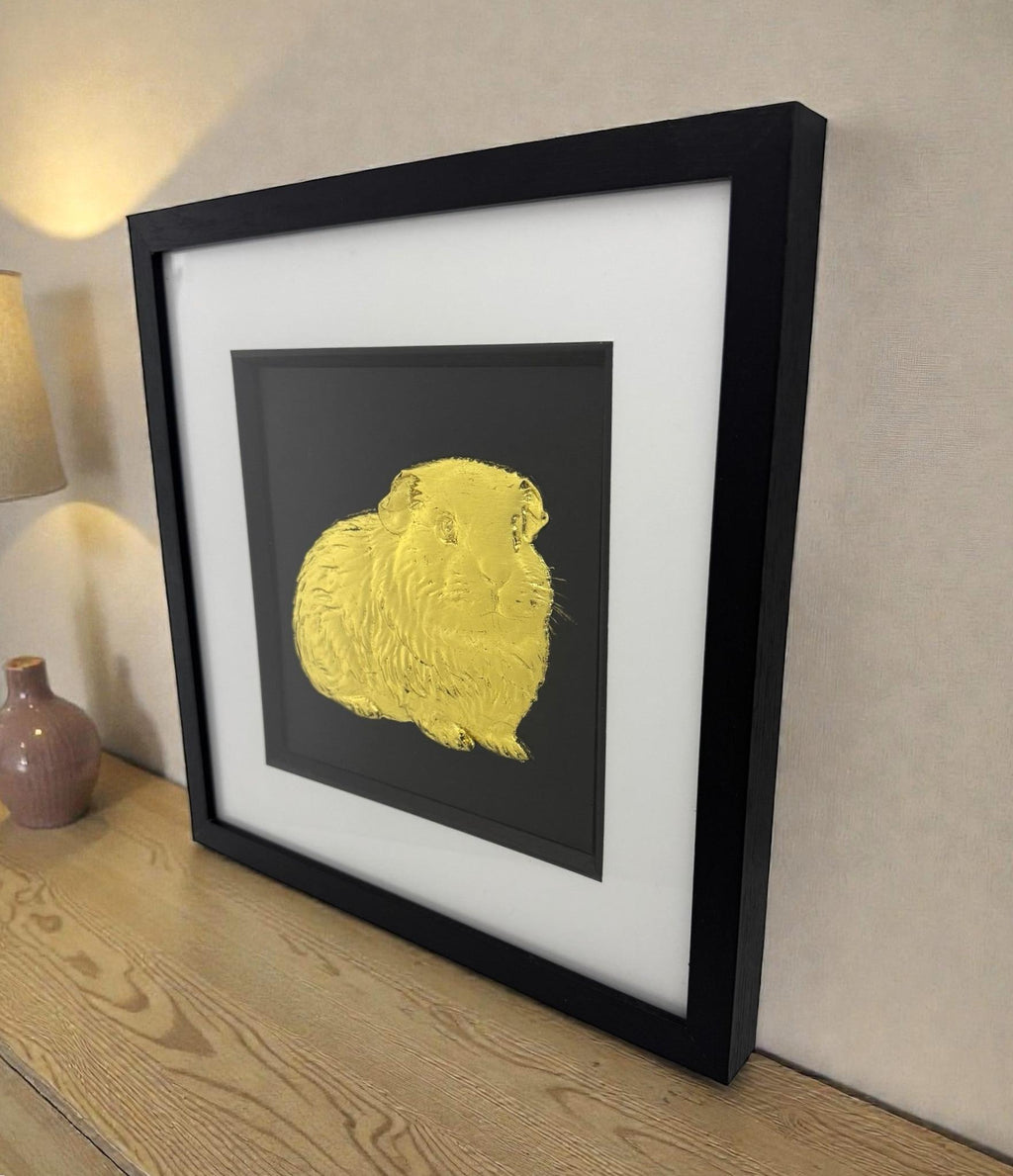 A gold leaf Guinea Pig framed art piece displayed on a wooden sideboard, showing the deep black box frame and premium finish suitable for home decor.
