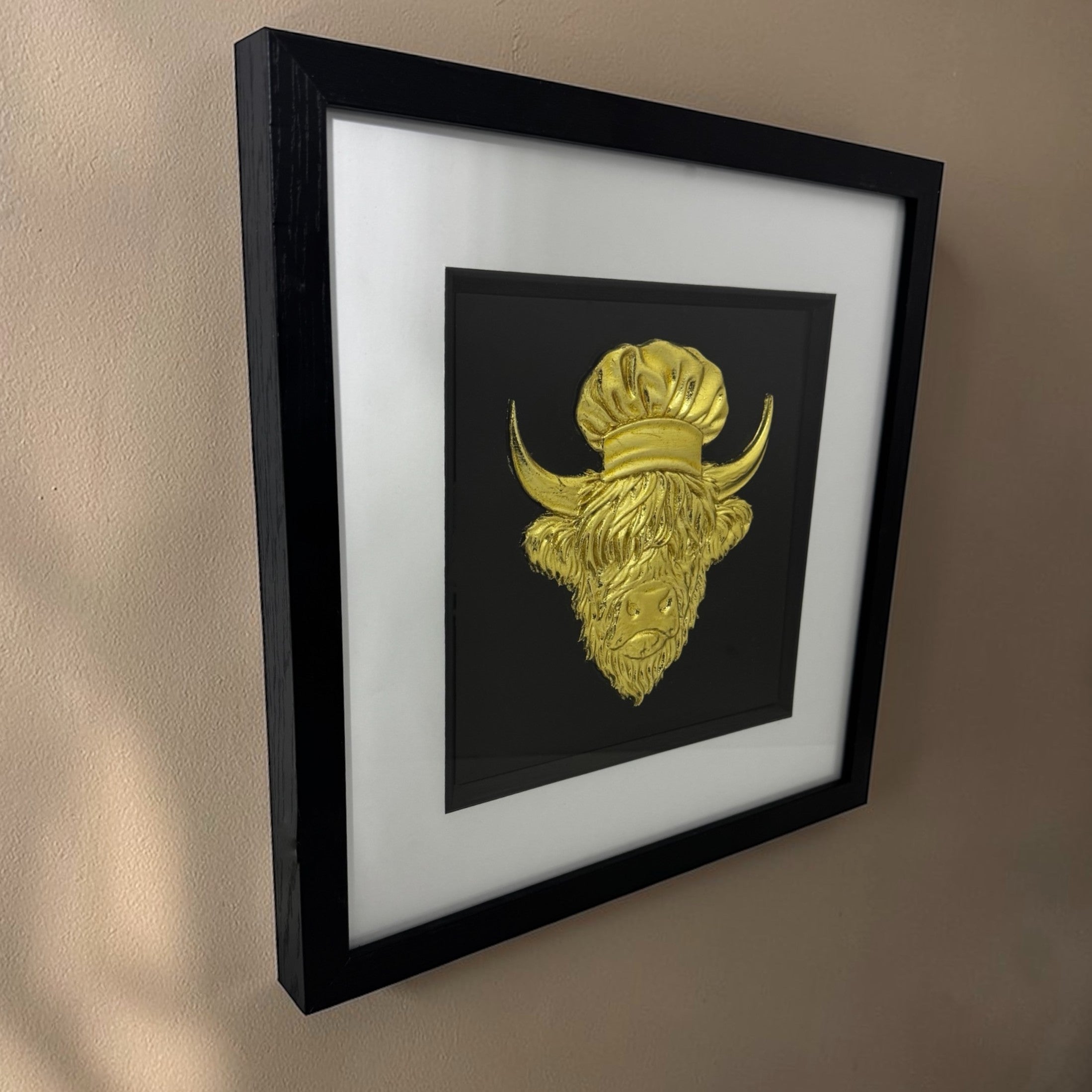Angled side view of a gold Highland cow chef relief in a black box frame hanging on a greige beige wall.