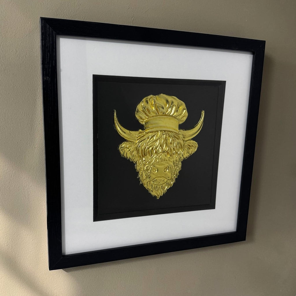 Angled view of a gold Highland cow chef relief in a black box frame hanging on a beige wall.