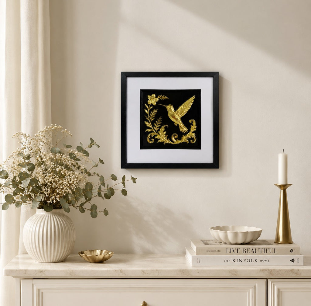 Gold hummingbird artwork styled above a cream console with vase, foliage, books and candle holder