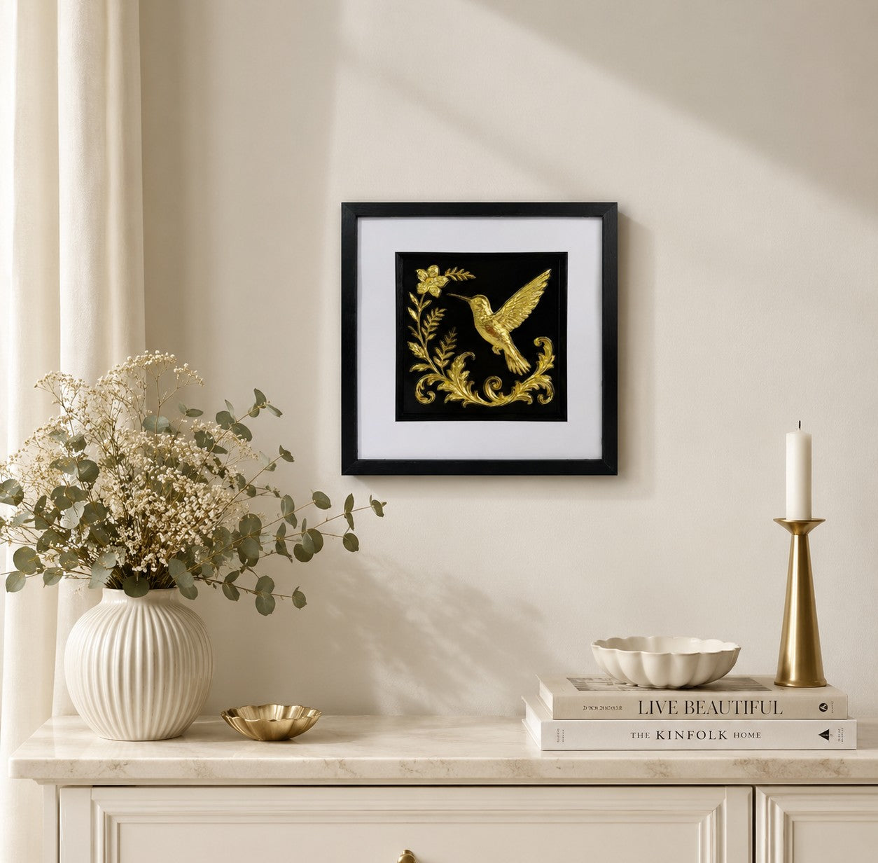 Gold hummingbird artwork styled above a cream console with vase, foliage, books and candle holder