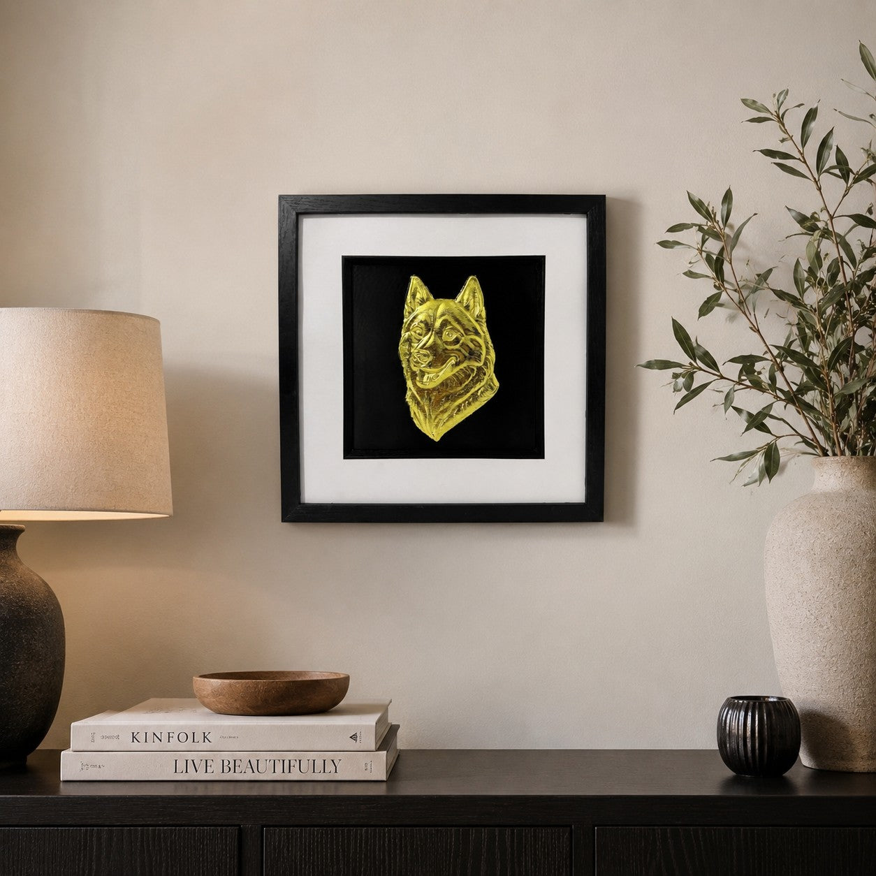 Framed gold husky dog wall art mounted on a neutral living room wall above a black sideboard with lamp, books and ceramic vase décor.
