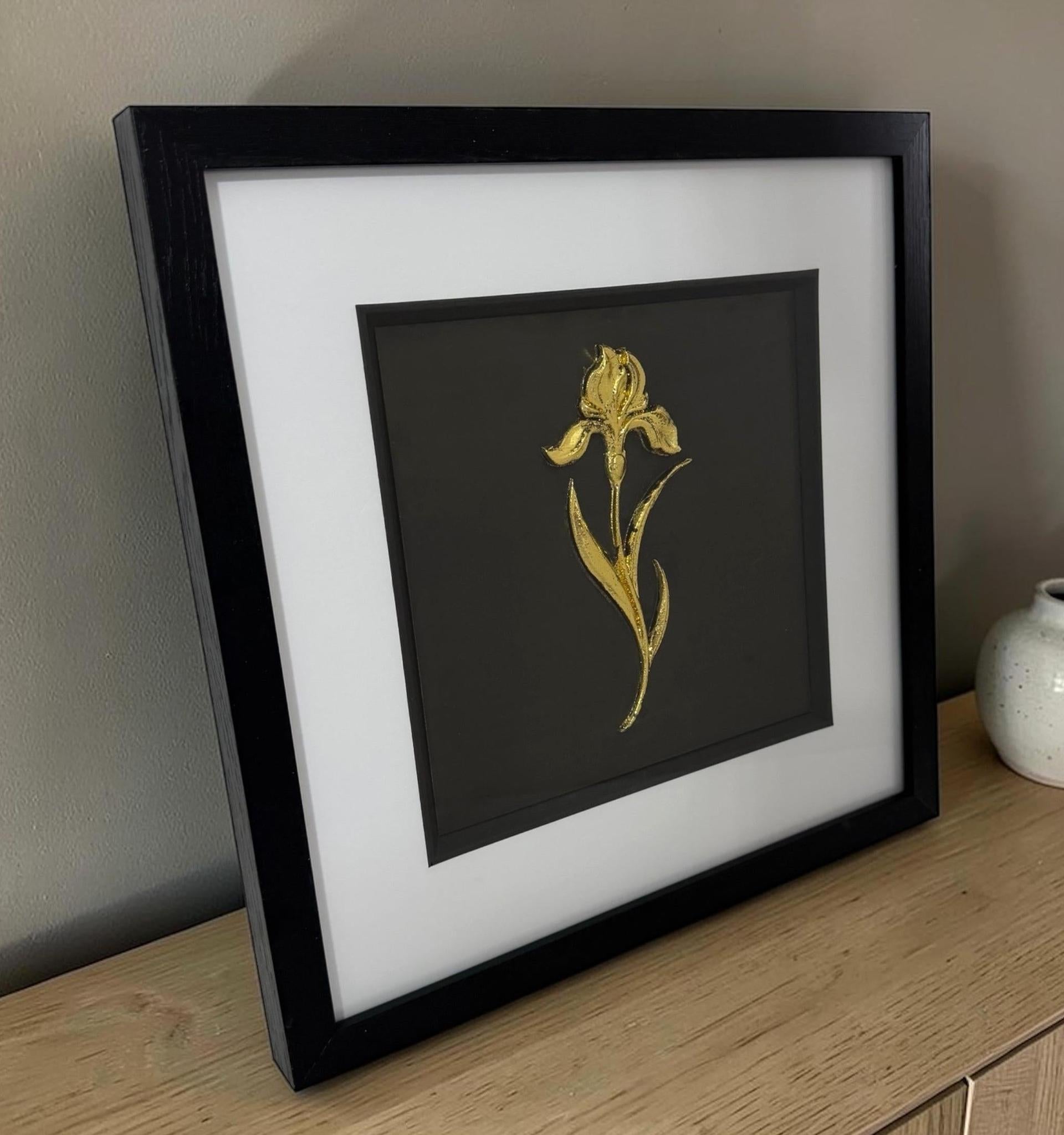 Gold iris raised relief in a black frame with white mount, styled on a wooden sideboard against a neutral wall.