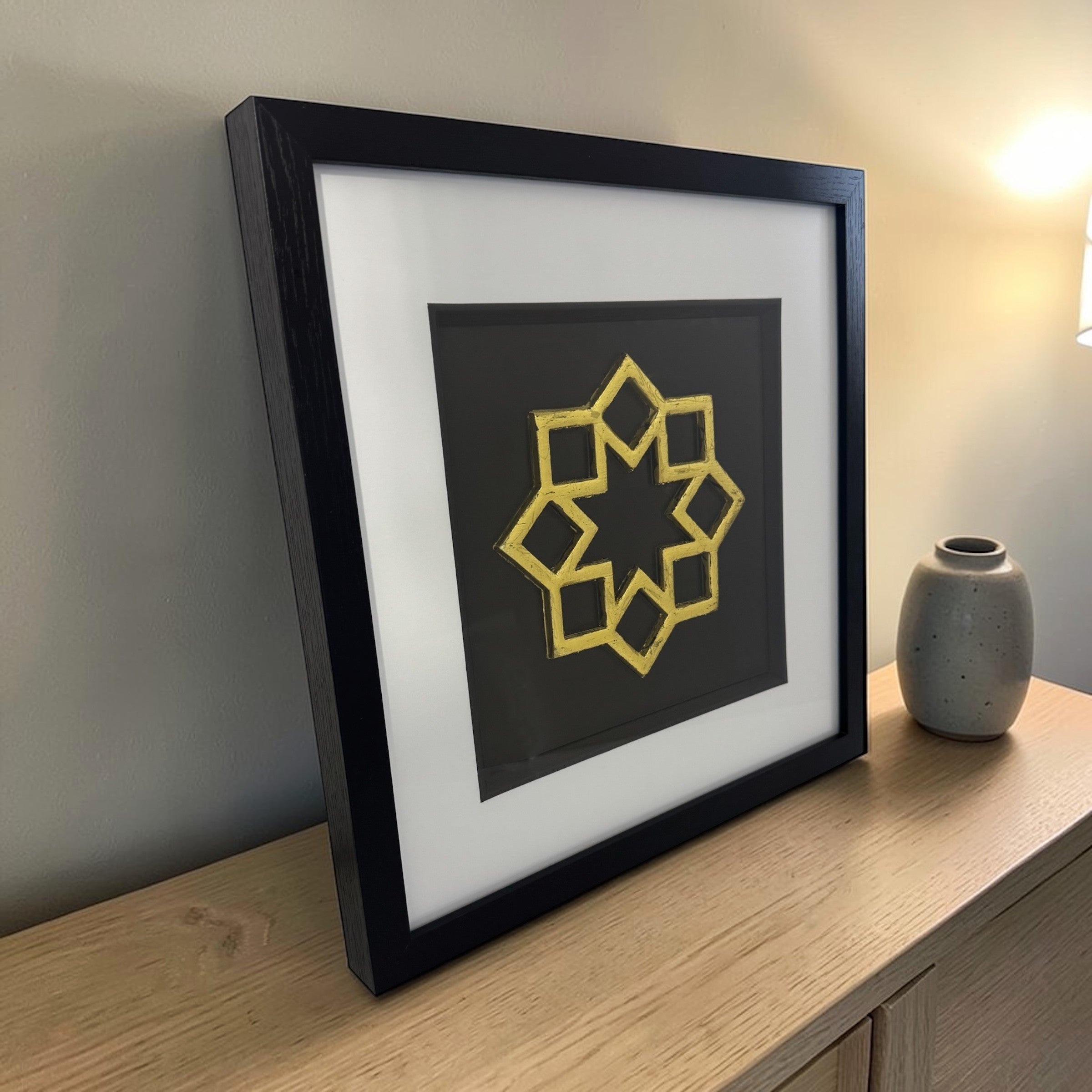 Angled view of the gold framed Islamic geometric star relief with glass front on a light wood sideboard beside a ceramic vase and lamp