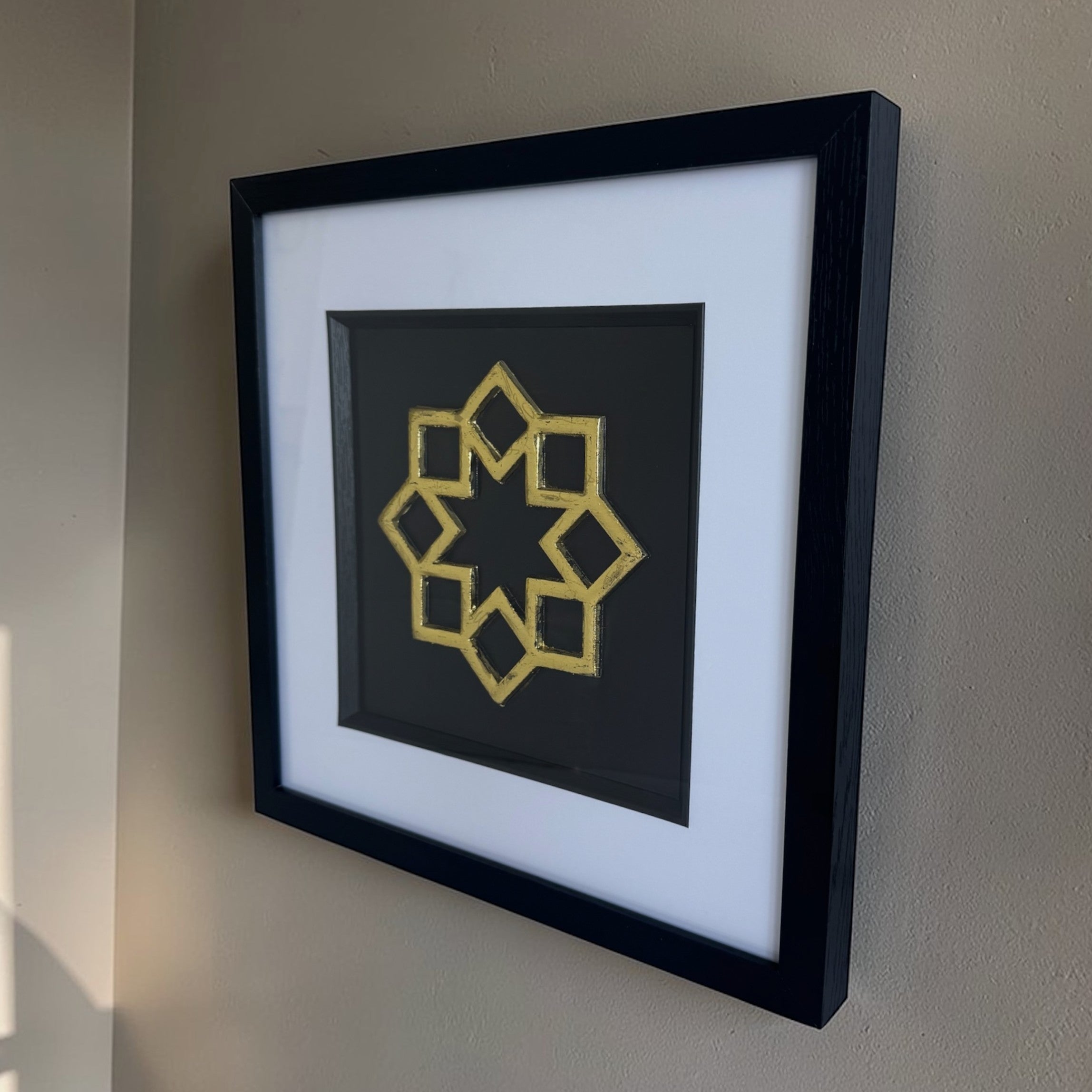 Angled gold Islamic 8 point star wall decor in a black frame with a white mount on a beige wall