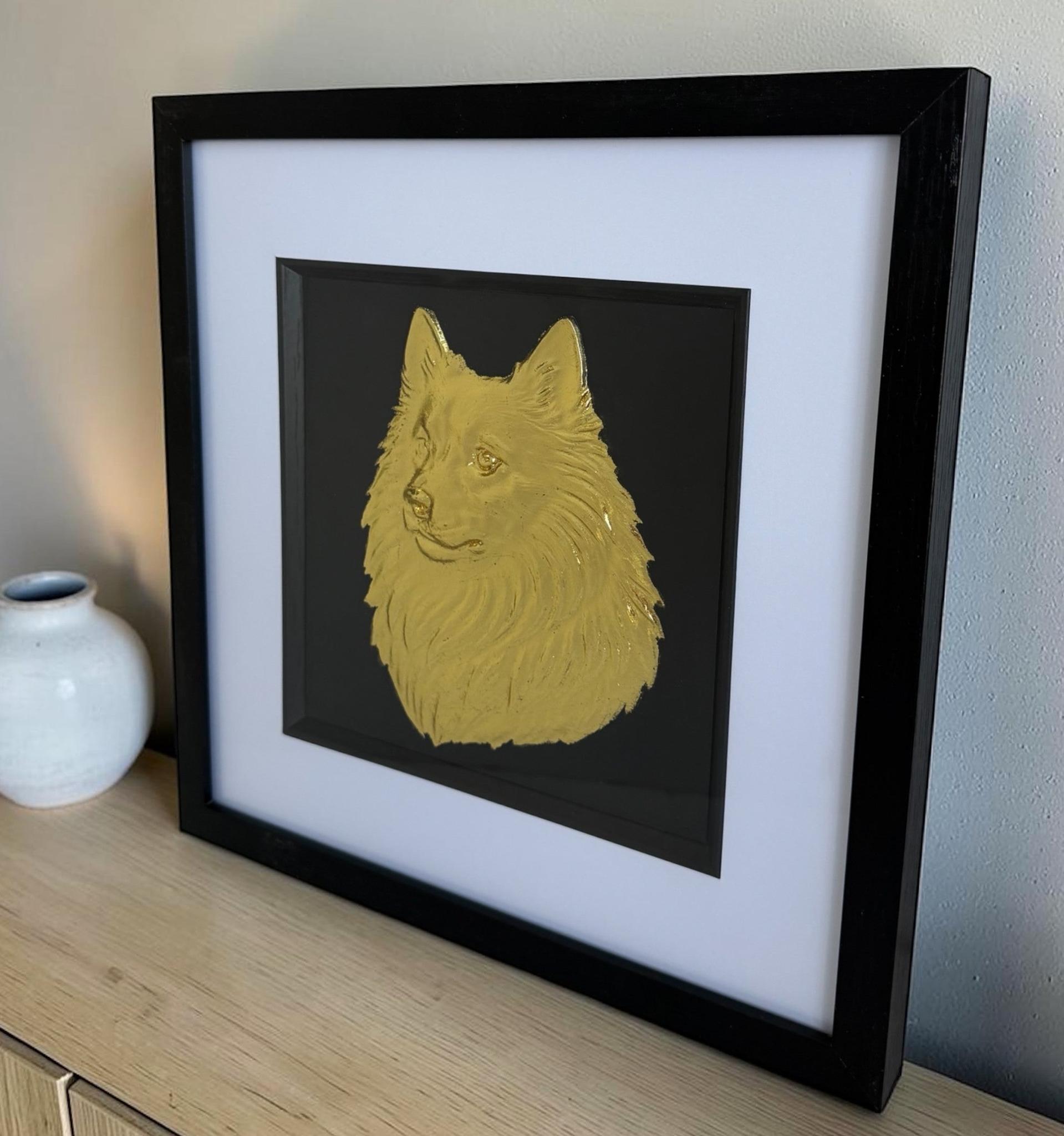 Gold Japanese Spitz relief portrait in a black box frame on a light wood surface beside a white ceramic vase.