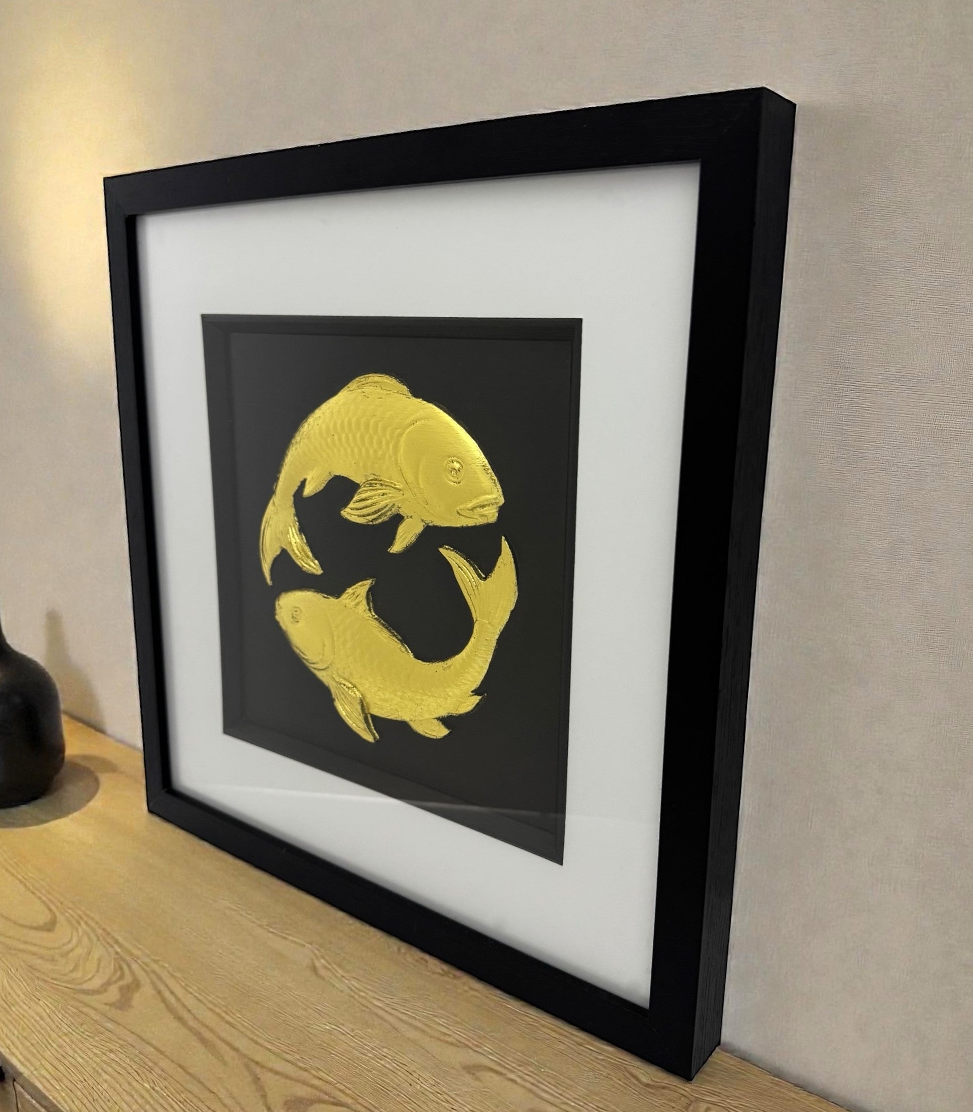 A gold leaf Koi Carp art piece displayed on a wooden sideboard, showing the 3D texture of the scales and the deep gallery frame.