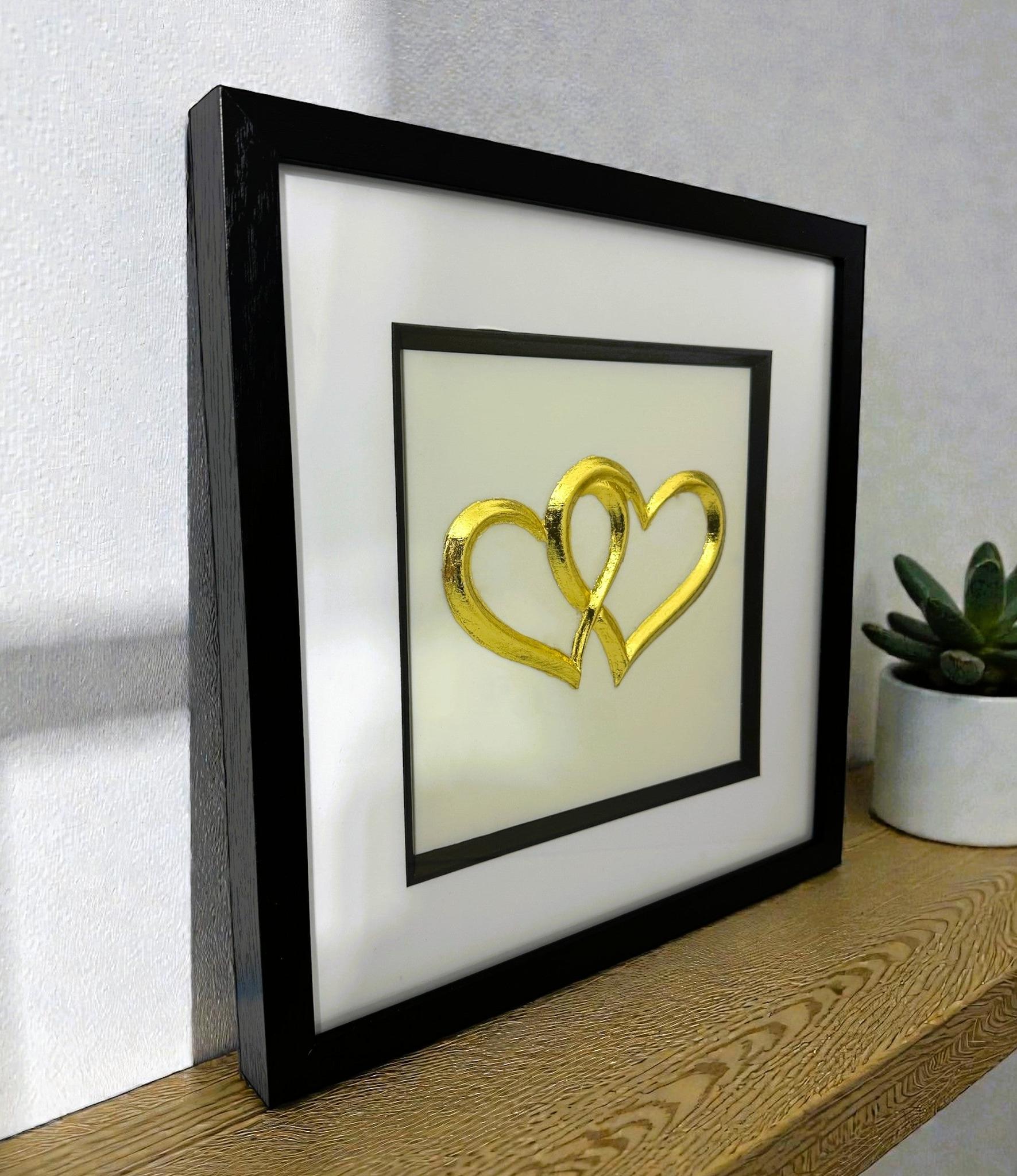 Angled desk view of gold leaf joined hearts wall art on a light background in a black wooden frame