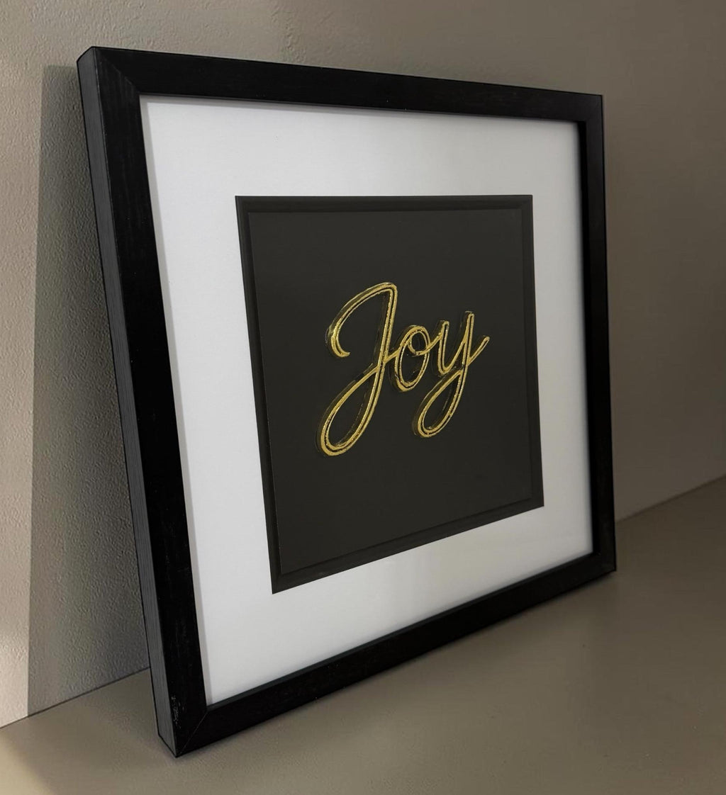 Angled gold Joy word artwork leaning on a neutral surface with black box frame and glass front.