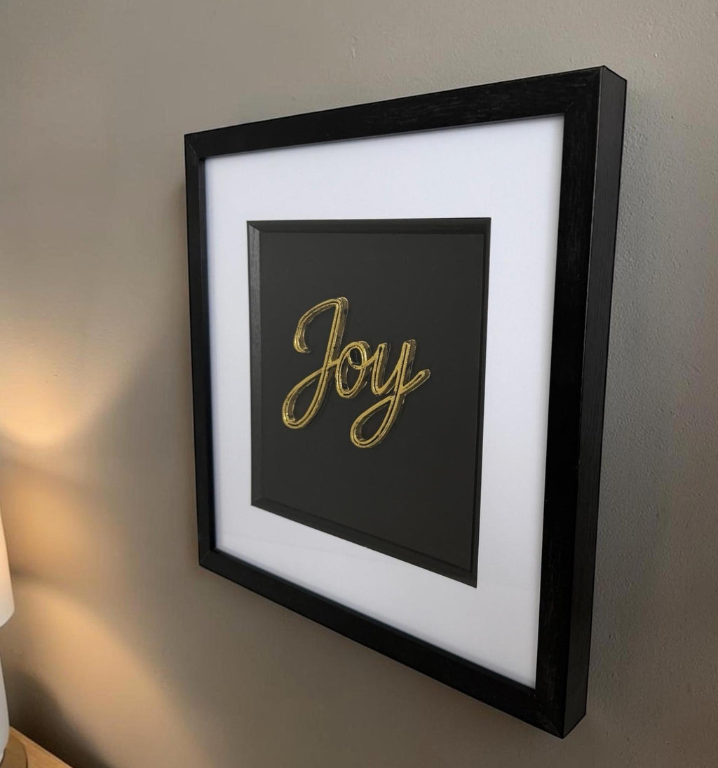 Gold Joy word artwork on a wall showing raised lettering, white mount, black centre and deep black frame.