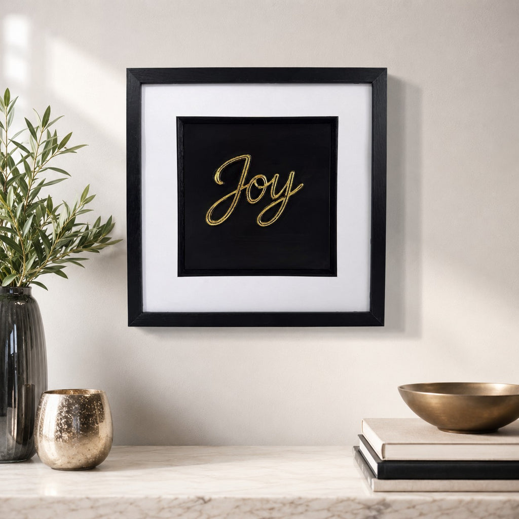 Gold framed Joy word artwork styled on a neutral wall with plant, books, bowl and black square frame.