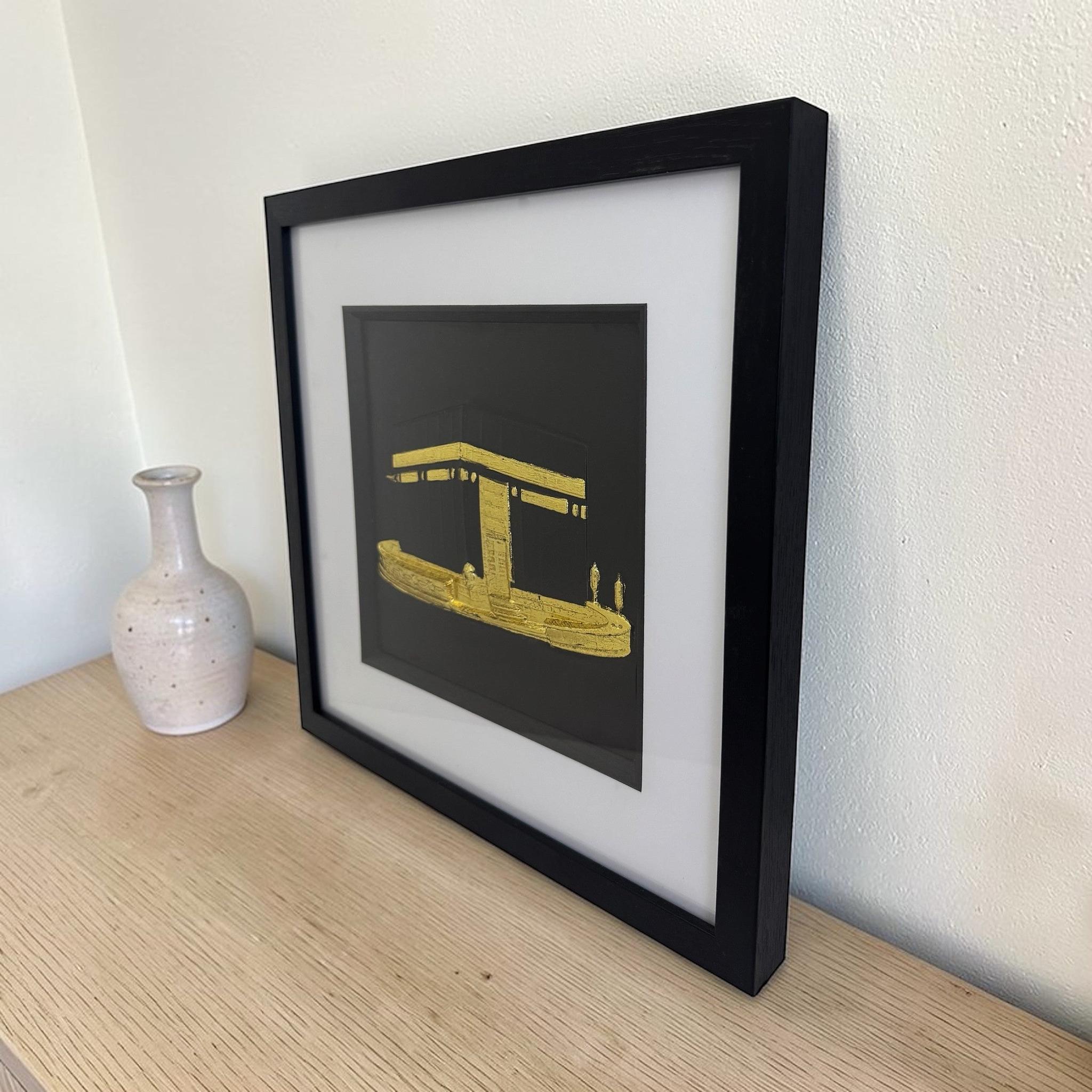 Gold Kaaba wall decor in a black box frame, styled on a wooden sideboard beside a ceramic vase.