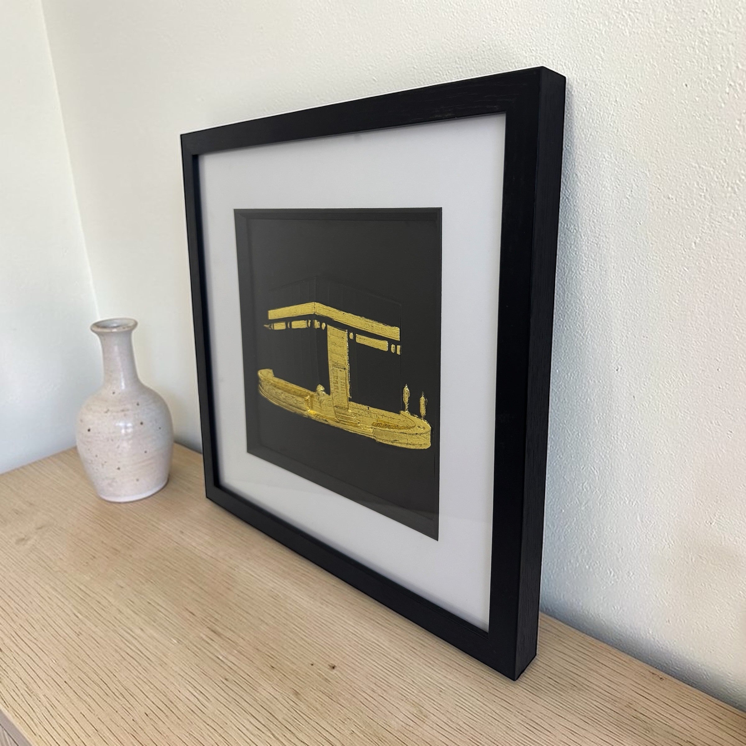 Gold Kaaba wall decor in a black box frame, styled on a wooden sideboard beside a ceramic vase.